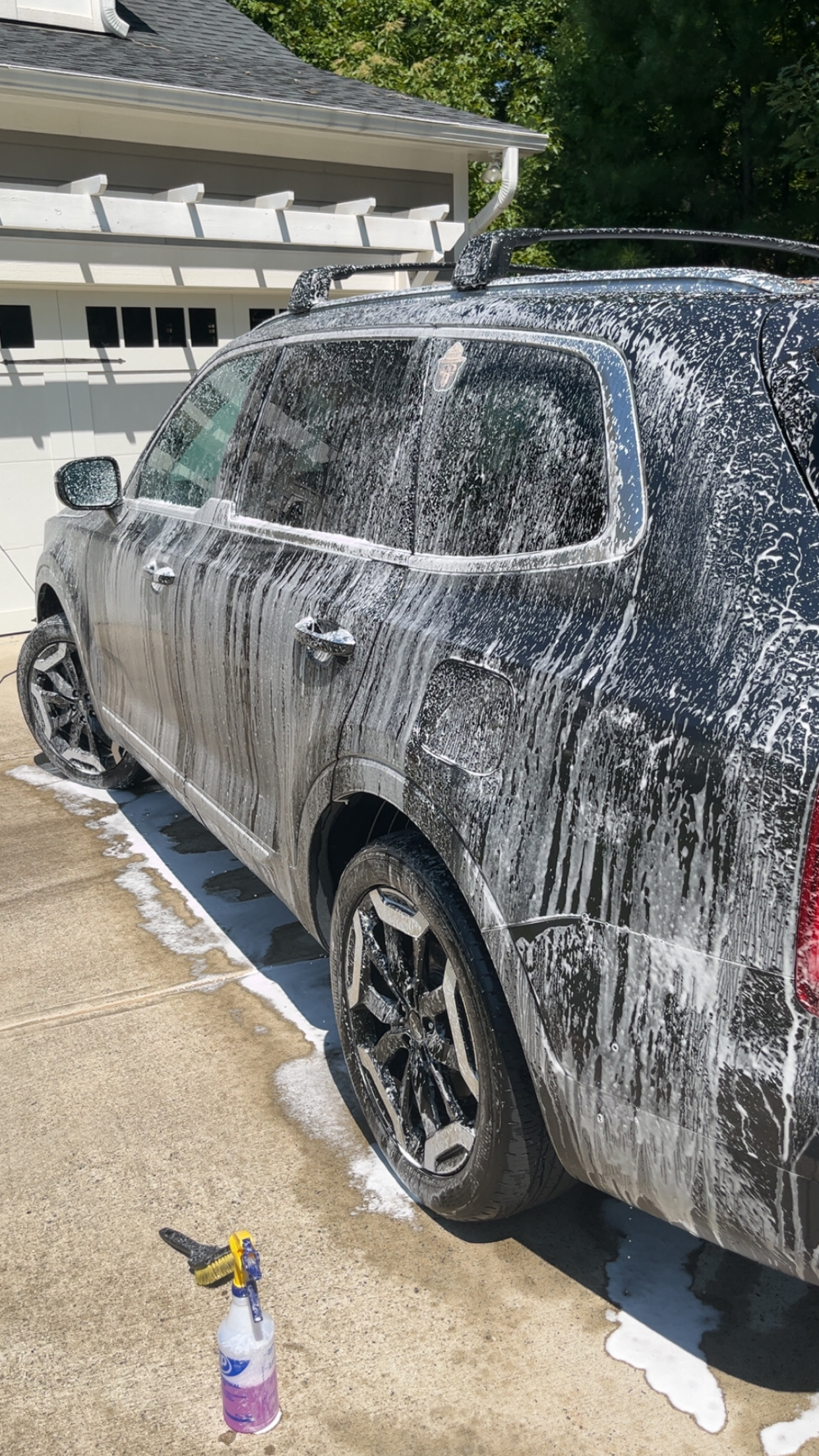 Dark SUV covered in thick foam suds mid-wash with a foam cannon spray bottle nearby